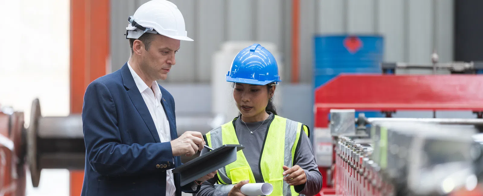 Engineer and worker in safety helmets discussing plans in industrial factory setting next to machinery.