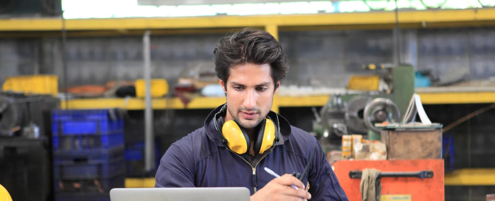 Male engineer with safety earmuffs around his neck working on a laptop in an industrial workshop.