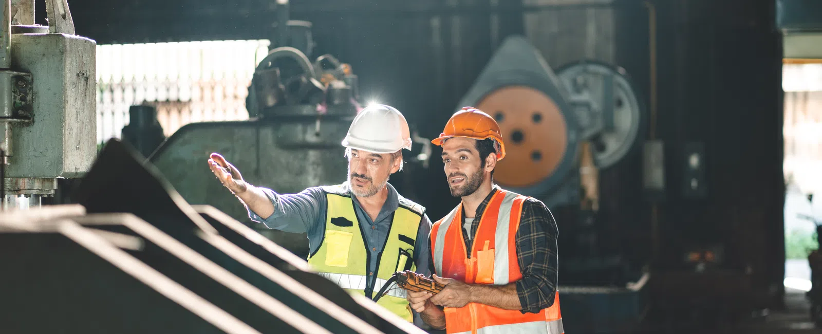 Two construction workers wearing helmets and safety vests discussing plans inside an industrial workshop.
