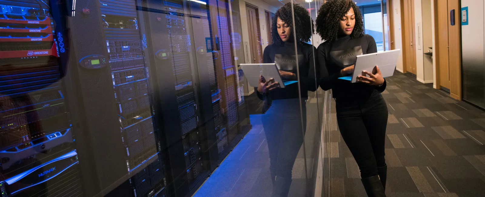 Woman in black working on laptop in server room hallway with glass wall reflection and blue lighting