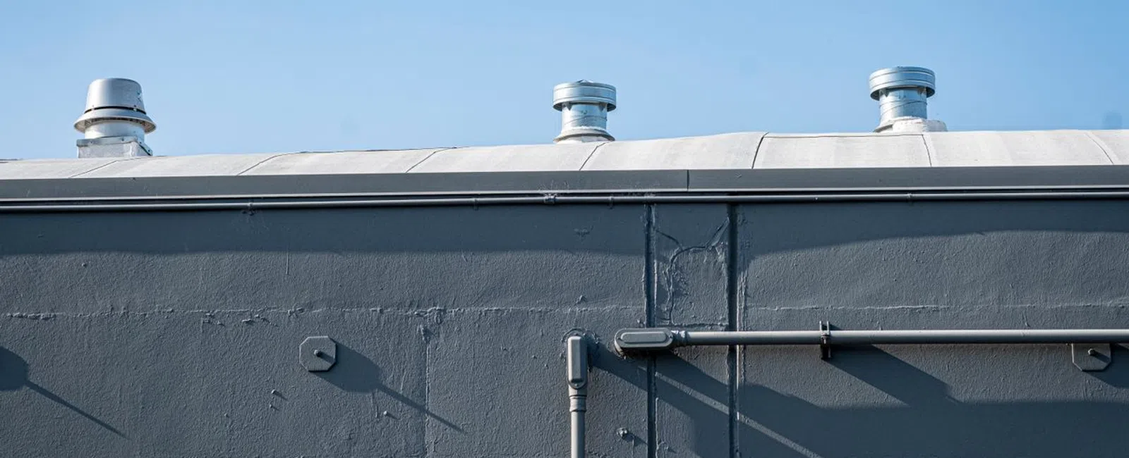 Metal rooftop vents on a gray industrial building wall under a clear blue sky with shadowed pipes and windows.