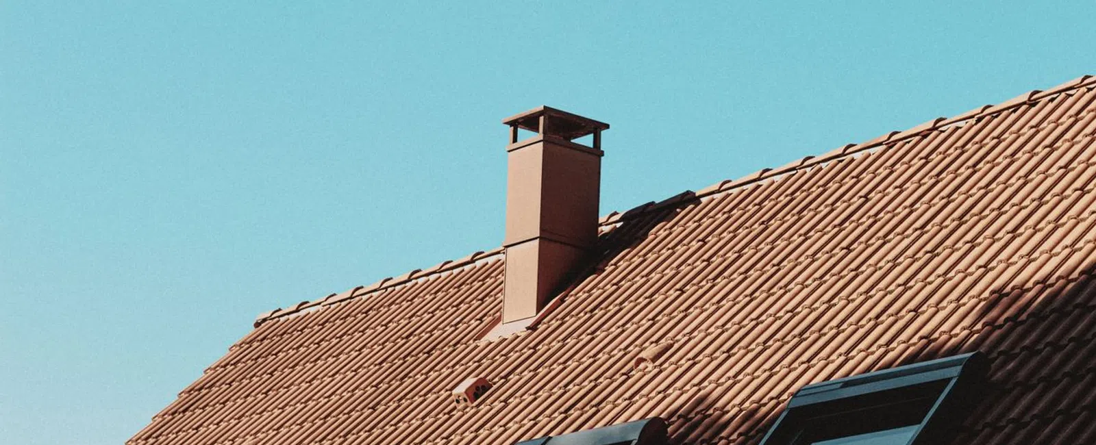 Brown tiled roof with skylights and chimney under clear blue sky in daylight.