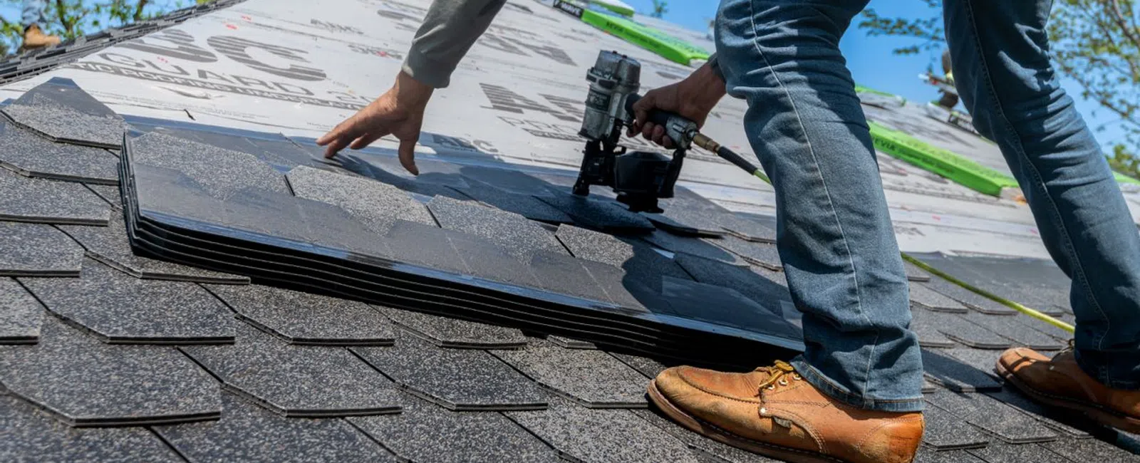 Worker installing asphalt roof shingles on a house using a nail gun on a sunny day.