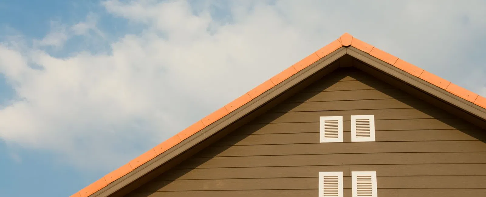 Brown house gable with orange roof tiles and four small white windows under a blue sky with clouds