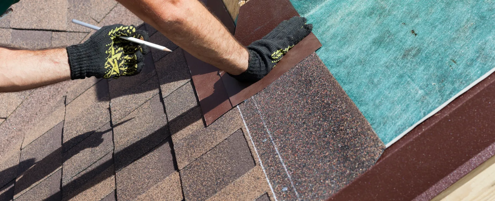 Close-up of a worker installing brown roof shingles with gloves and a pencil marking alignment on a sunny day.