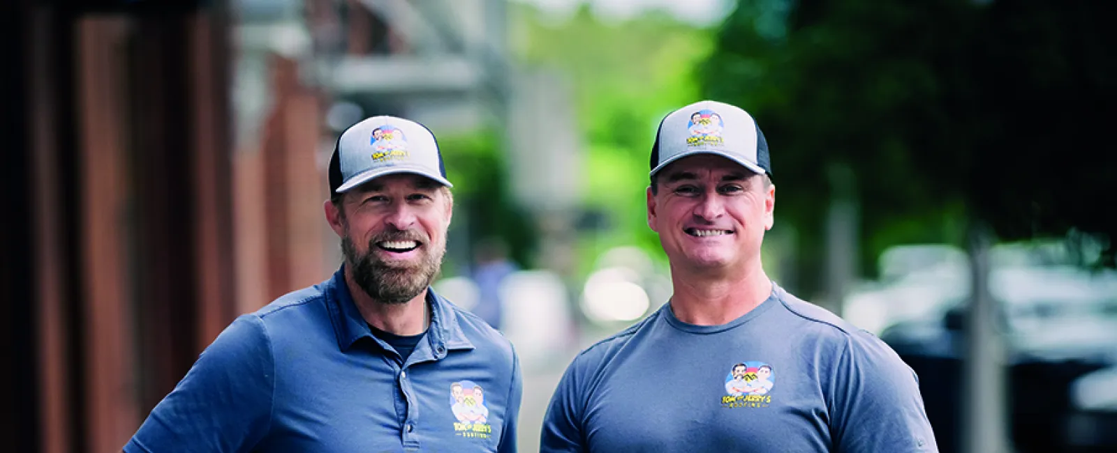 Two men wearing matching blue company shirts and hats standing on a city sidewalk smiling at the camera