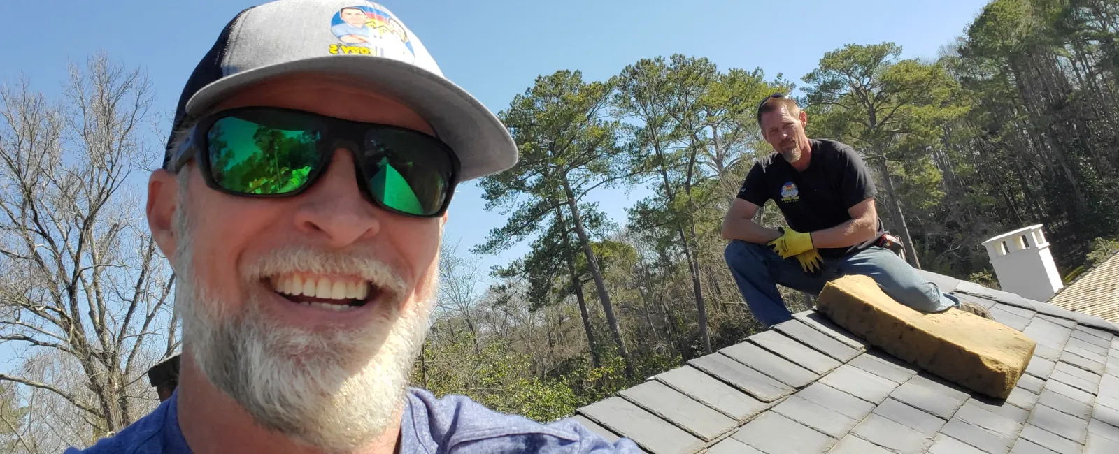 Two men working on a gray shingled roof on a sunny day with trees in the background.