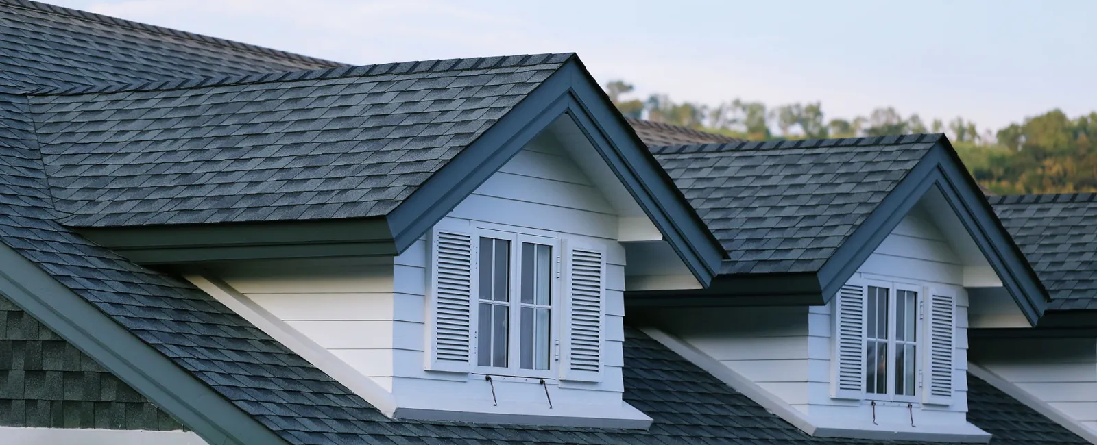 Close-up of house roof with dark gray shingles and two white dormer windows with shutters