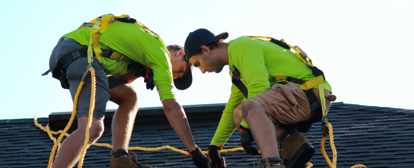 Two roofers in safety harnesses measuring on a shingled roof under clear sky.