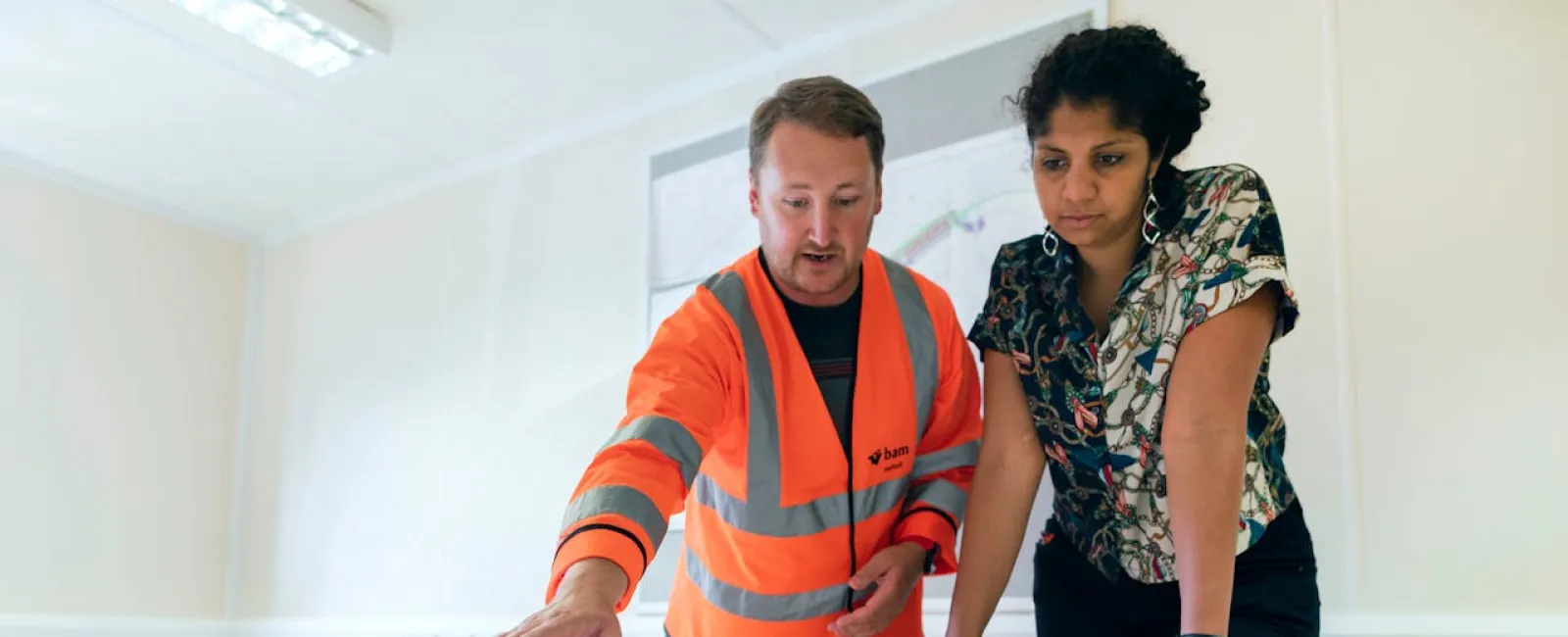 Man in orange safety jacket and woman discuss architectural plans on a table in a bright office.