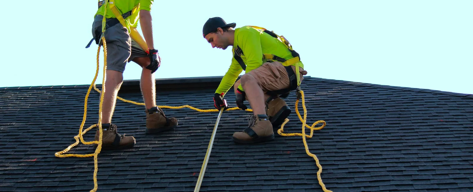 Two roofers in safety harnesses measuring and inspecting a shingled roof under clear daylight sky.