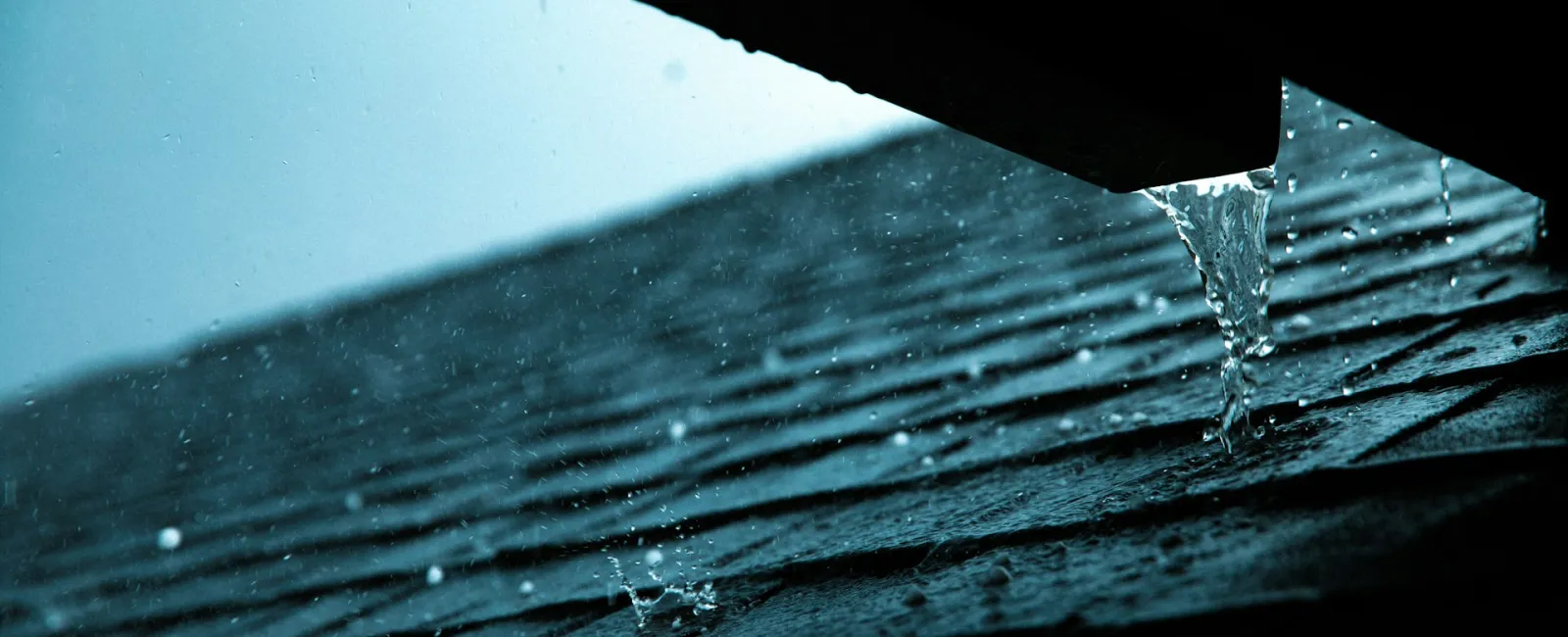 Water dripping from a roof edge onto dark shingles during rainfall in low light conditions.