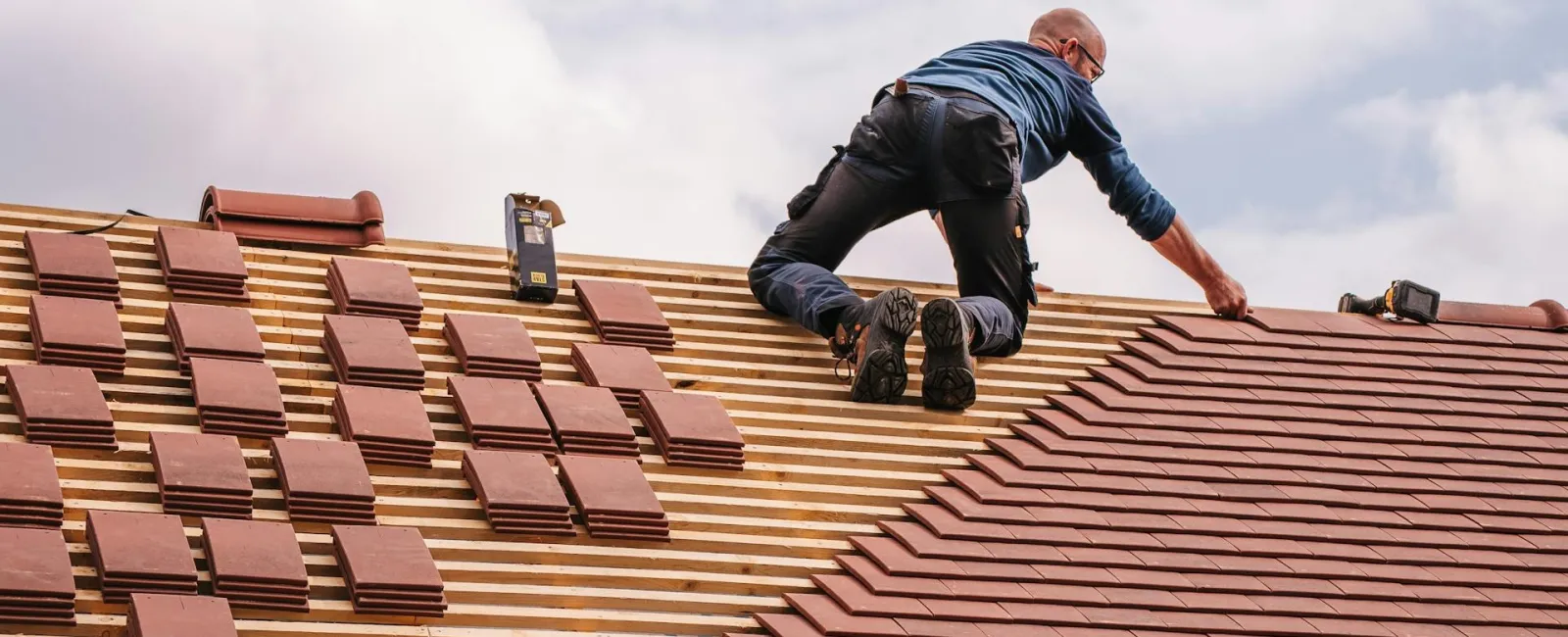 Roofer installing brown roof tiles on wooden framework against cloudy sky background.