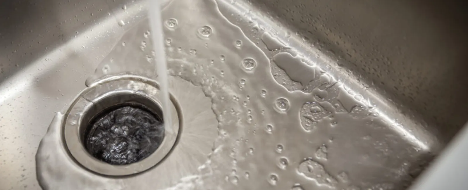 Water flowing into a stainless steel sink drain with bubbles and ripples visible around the drain area.