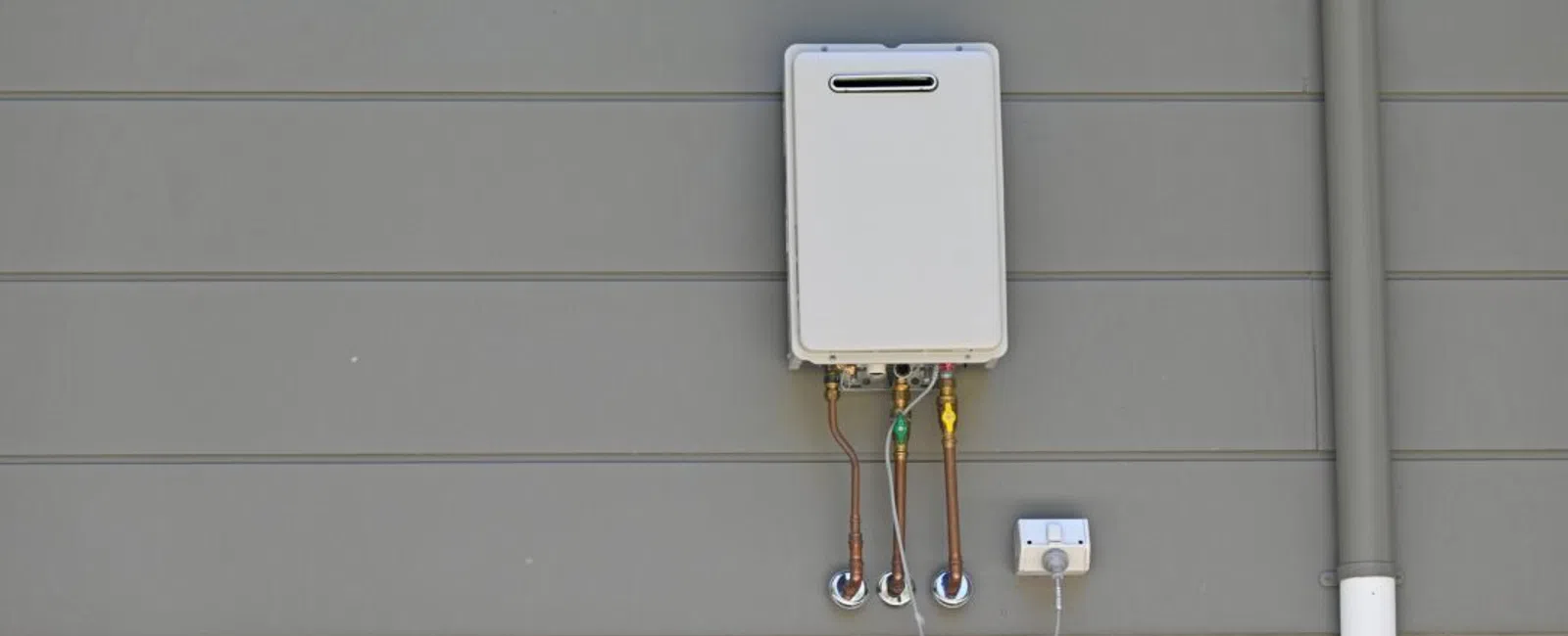 Wall-mounted tankless water heater installed on gray horizontal siding with connected pipes and power outlet.