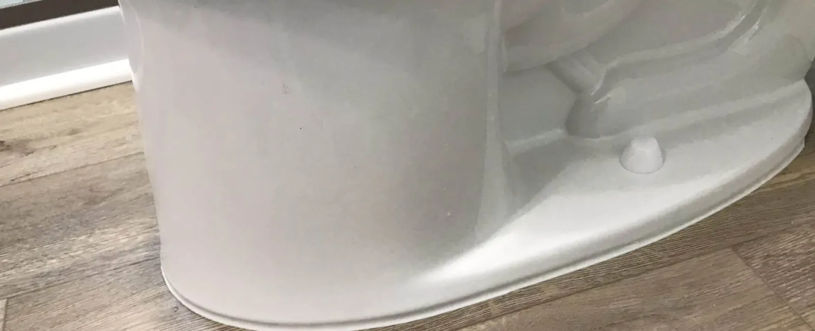 Close-up of white toilet base installed on wood-patterned vinyl flooring in bathroom corner.