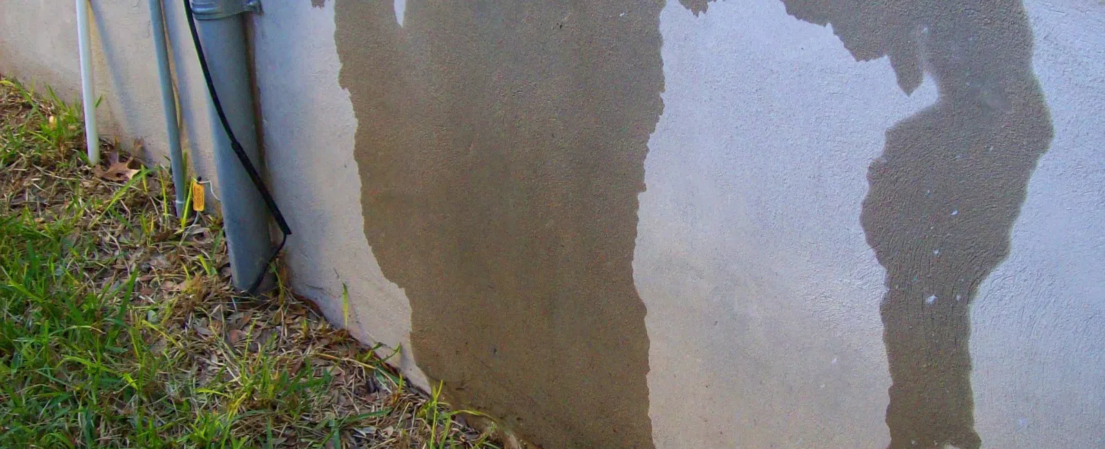 Water stains on a home's concrete foundation wall with adjacent grass and utility pipes visible.