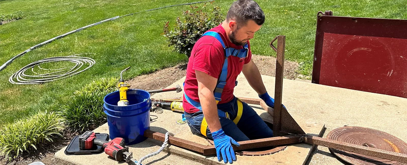 A worker in a red shirt repairs a sewer system, focused on the task with tools nearby.