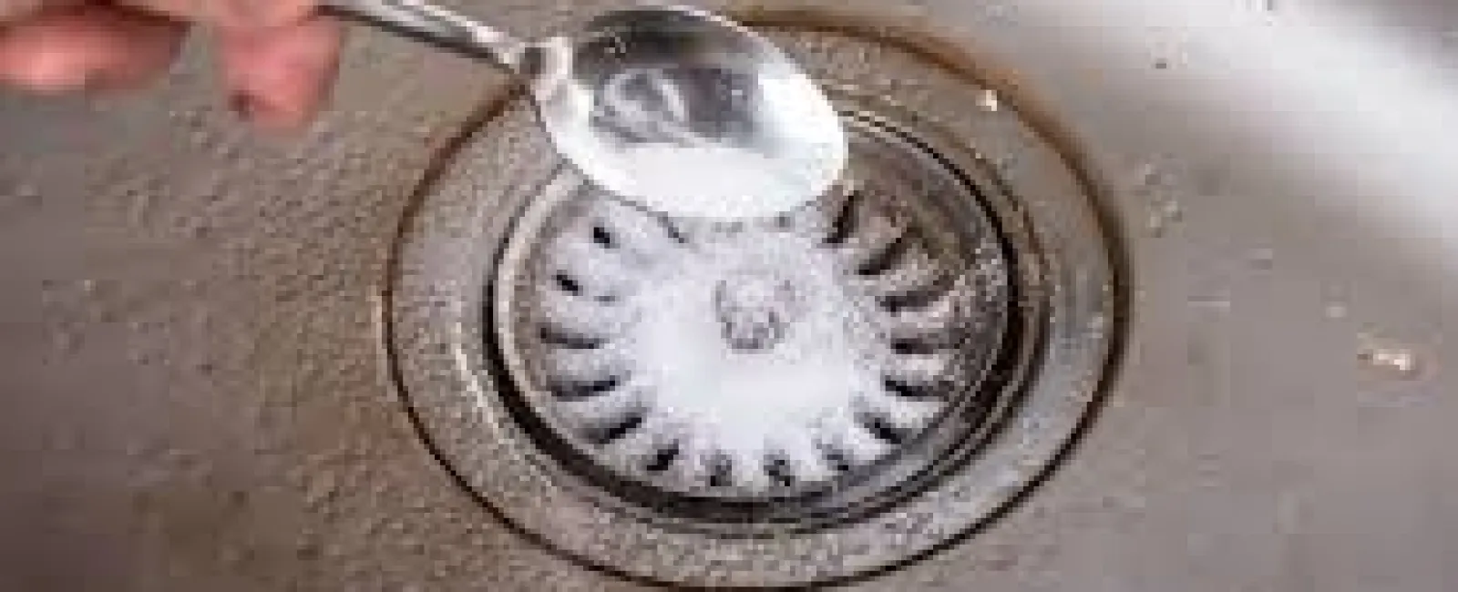 Hand pouring baking soda from a spoon into a kitchen sink drain for cleaning.