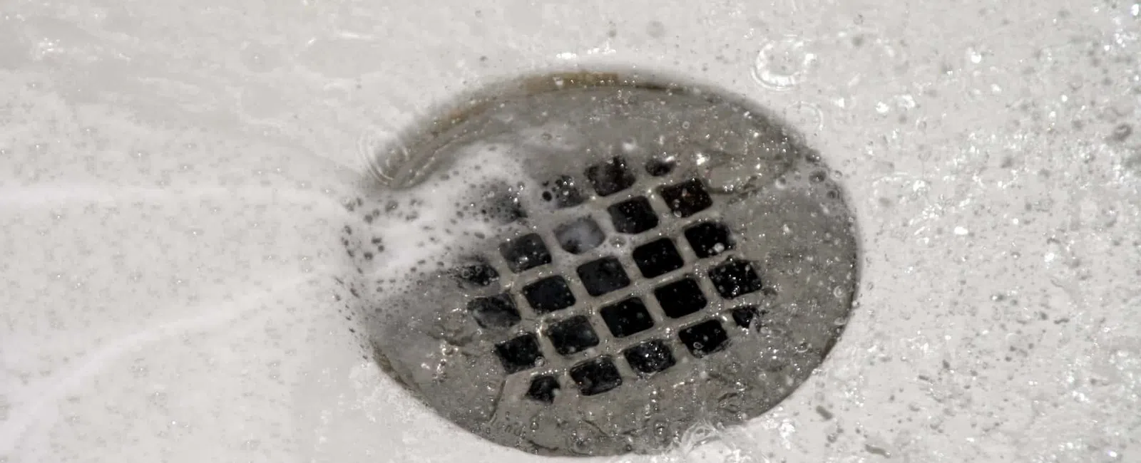 Close-up of a metal sink drain with water and soap bubbles flowing around it in a white basin.