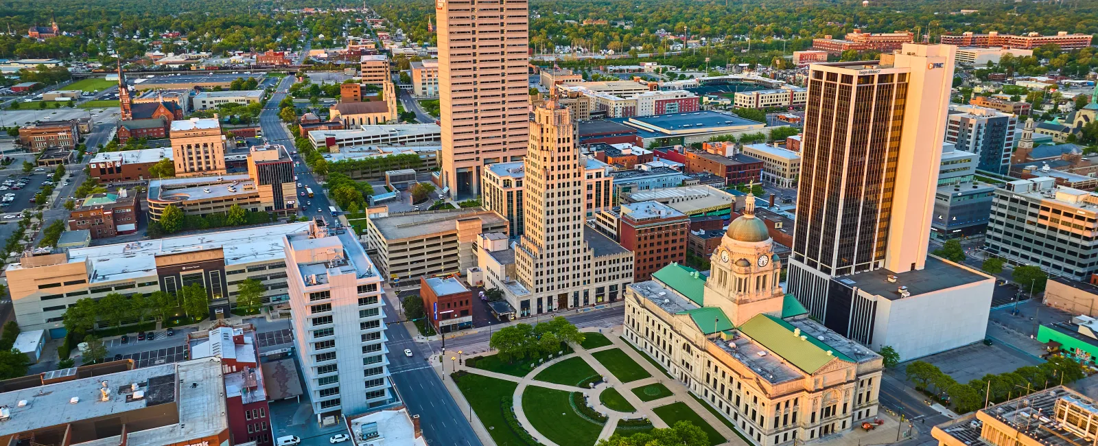 Aerial view of downtown city buildings with a historic courthouse and green park in the foreground at sunset.