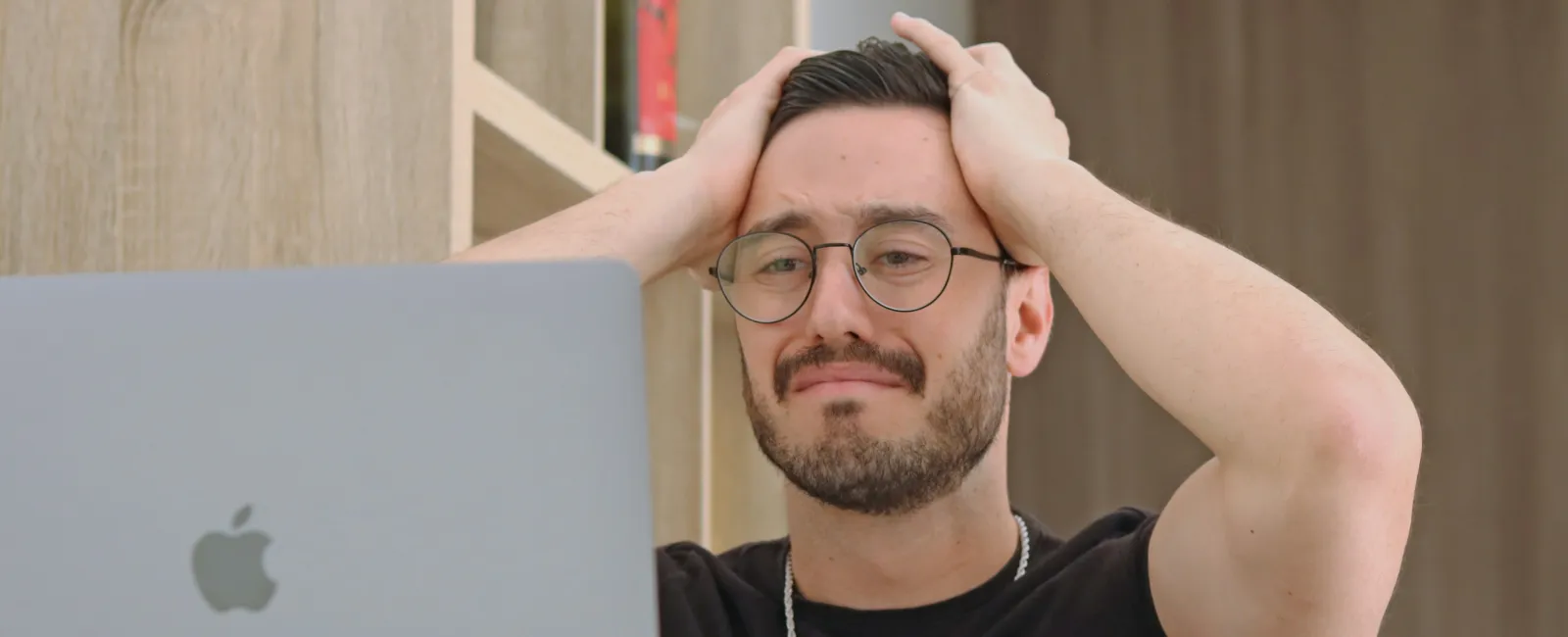 Frustrated man with glasses using MacBook laptop, holding head in stress after making small business IT mistakes.