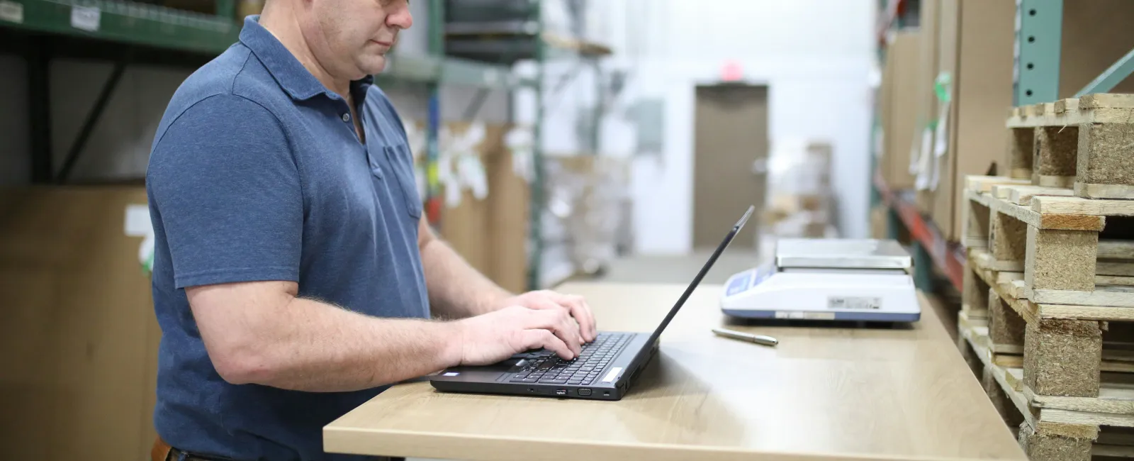 Man in blue shirt working on a laptop at a wooden table in a manufacturing warehouse with shelves and pallets.