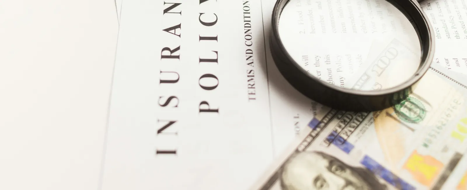 Magnifying glass on a cyber insurance policy document with US hundred-dollar bills partially visible beneath it.