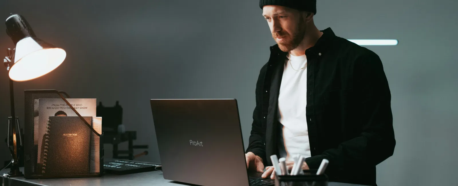 Man wearing beanie and black jacket doing cybercrime on a laptop at a metal desk under a desk lamp.