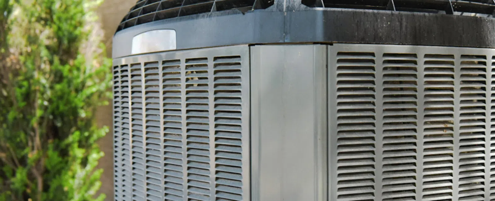 Close-up of an outdoor air conditioning unit with metal vents and protective grille beside greenery.