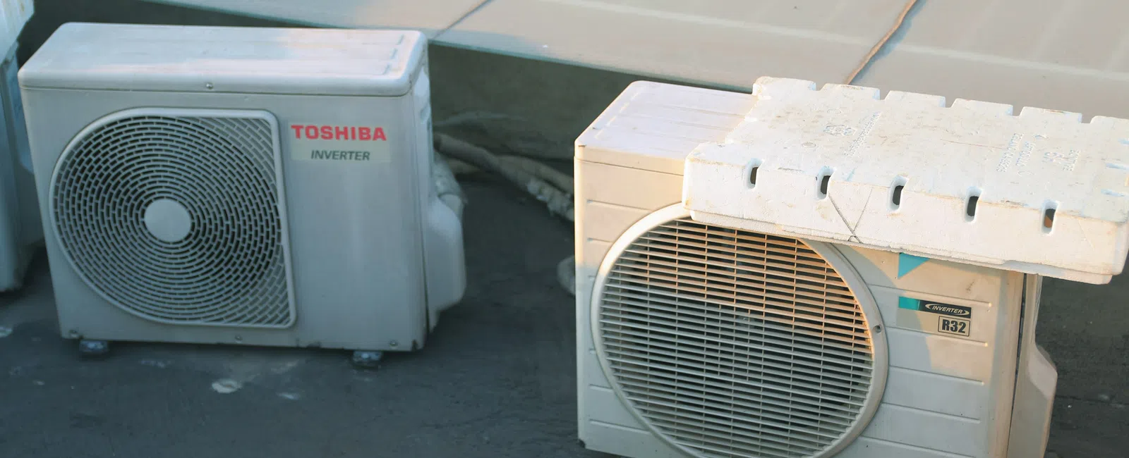 Two outdoor air conditioning units placed on a flat rooftop under sunlight with protective covering on one unit.