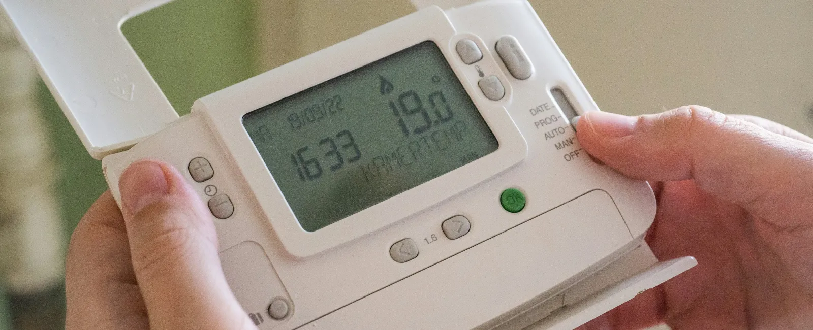 Hands adjusting a white digital thermostat displaying temperature and time settings indoors.