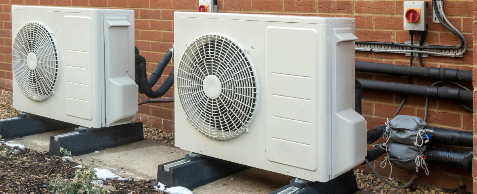 Two large outdoor air conditioning units installed on concrete slabs next to a brick wall with piping and wiring.
