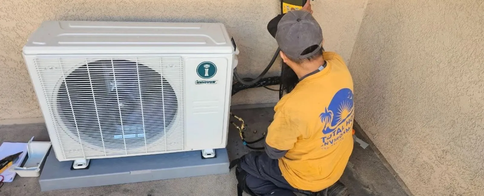 Technician installing or repairing an outdoor air conditioning unit on a concrete surface near a stucco wall.