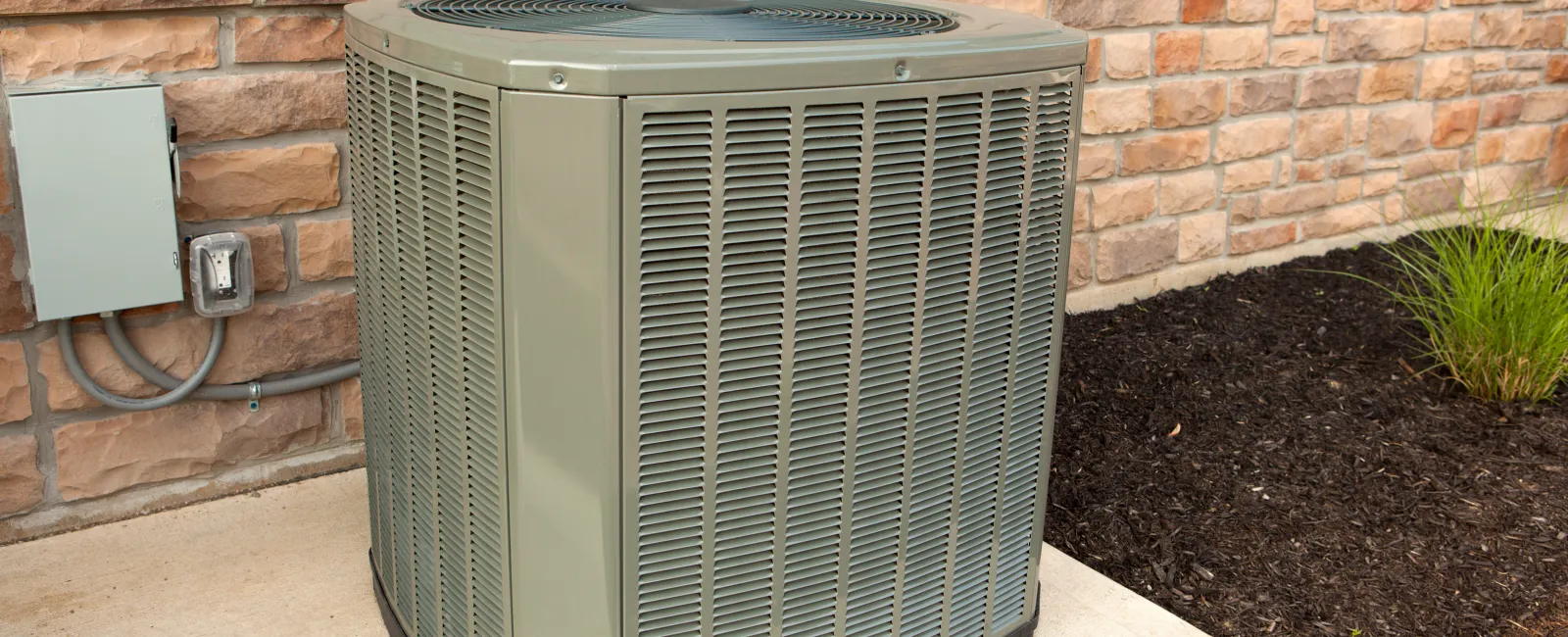 Outdoor air conditioning unit installed next to a house with brick siding and mulched garden bed