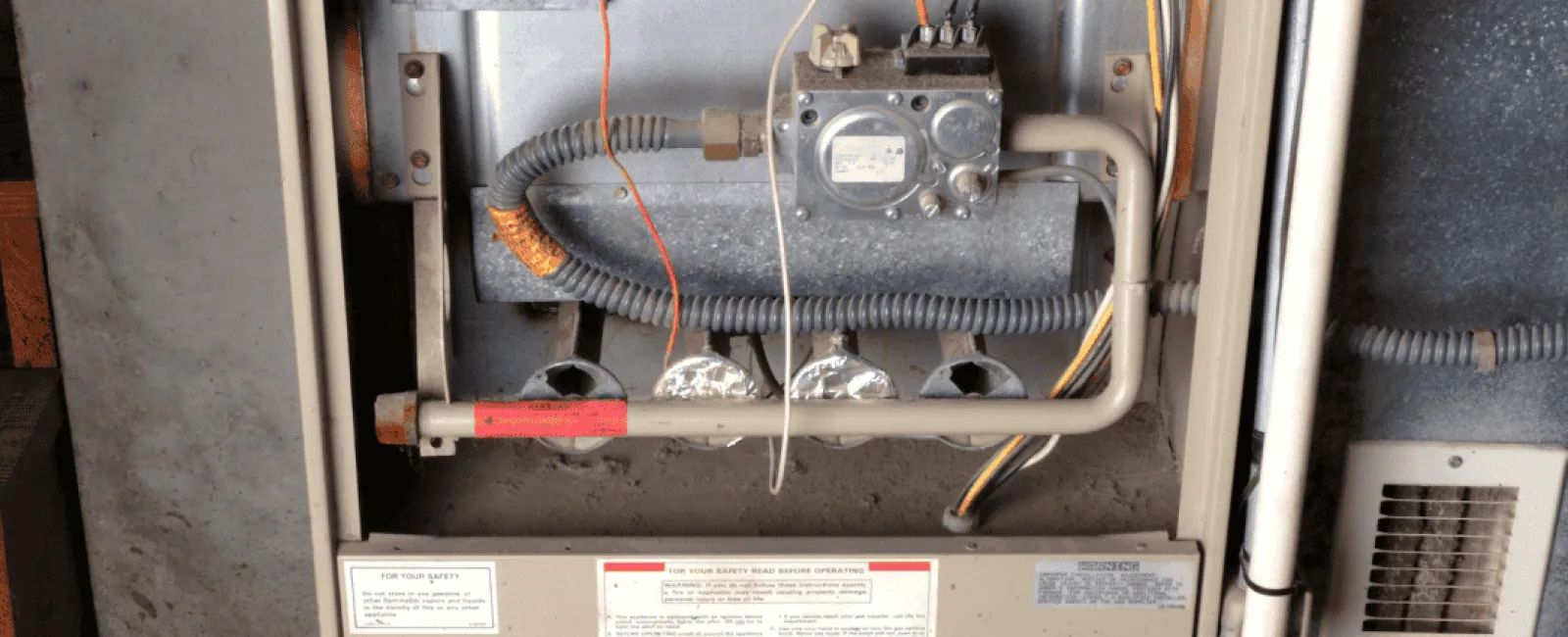 Old beige furnace with open panel showing gas valves, wires, and a vent pipe in a basement setting.