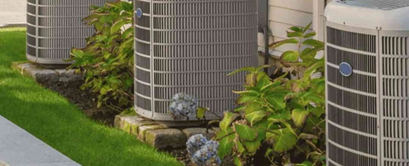 Three outdoor air conditioning units beside a house with green plants and flowers in a landscaped garden bed.