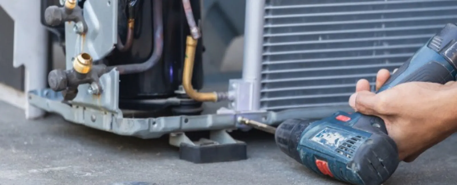 Technician using a cordless drill to repair an HVAC unit on the ground with tools nearby in focus.