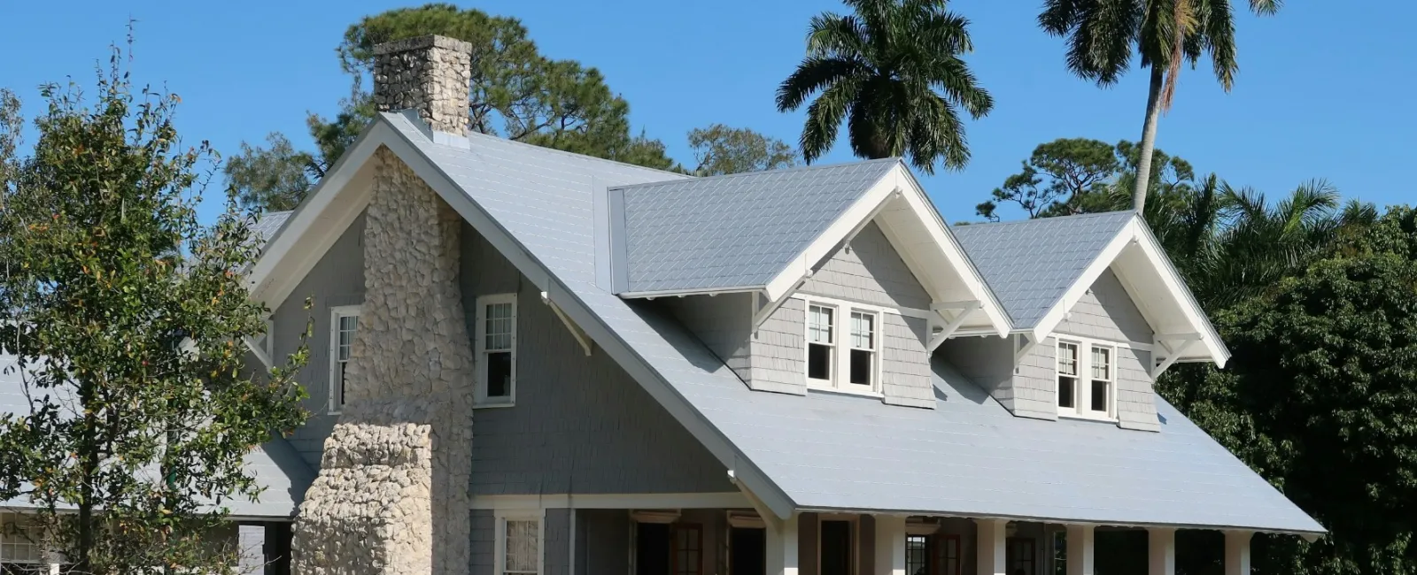 Gray craftsman-style house with stone chimney, front porch, surrounded by trees and palm trees under clear sky