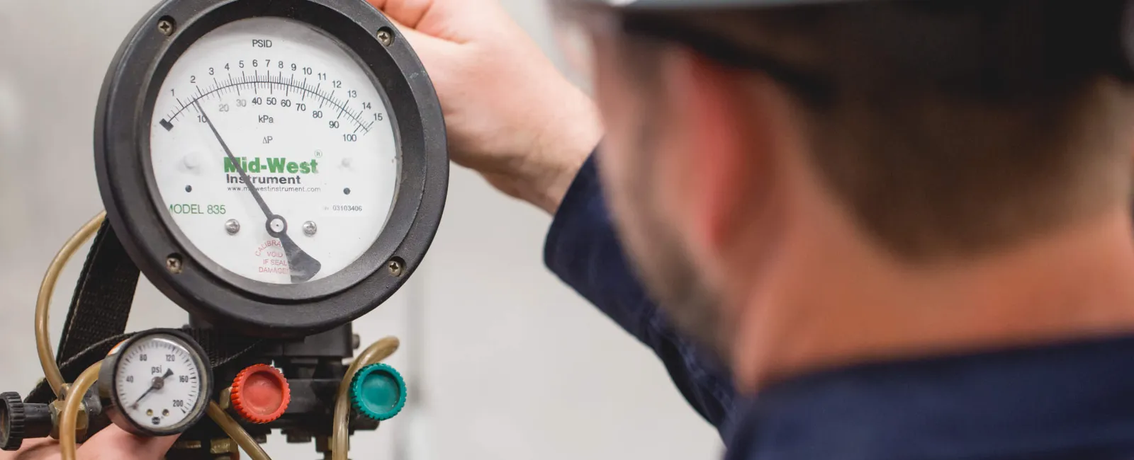 Technician in a hard hat adjusting Mid-West Instrument pressure gauge for HVAC system maintenance.