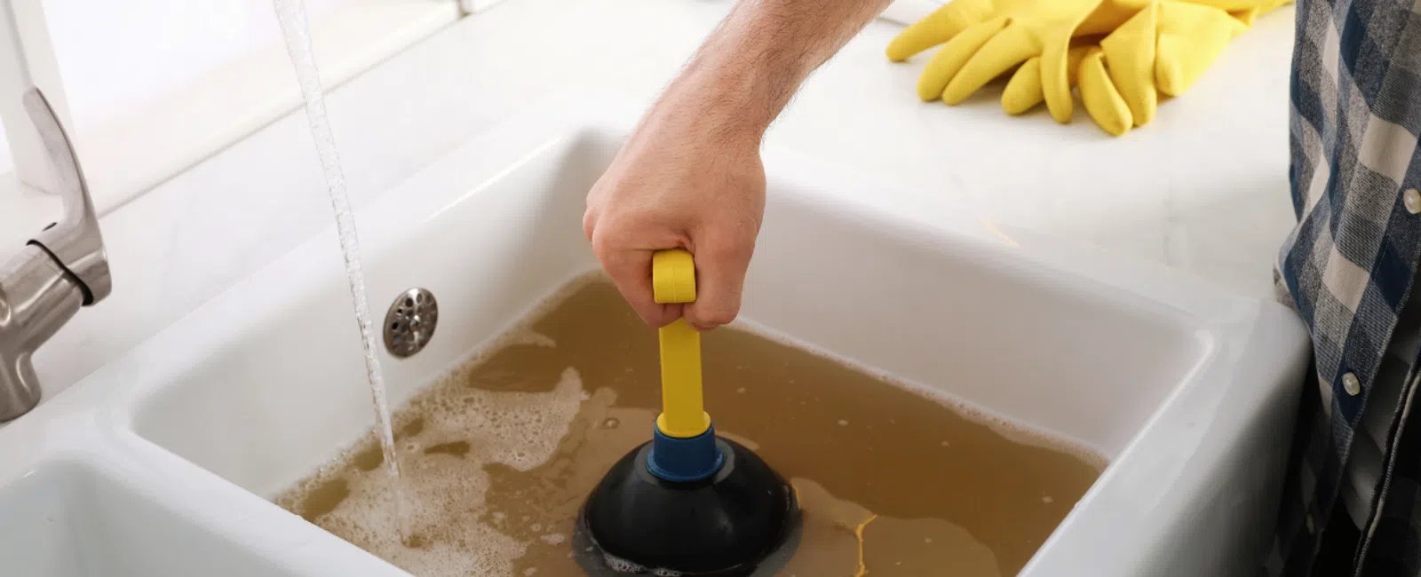 Hand using plunger to clear clogged brown water in kitchen sink with running faucet and yellow gloves nearby