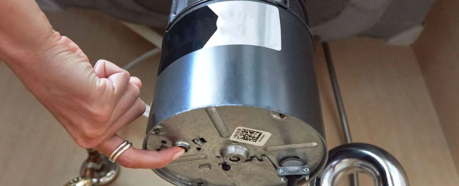 Hand adjusting the bottom of a garbage disposal unit installed under a kitchen sink with metal pipes visible.