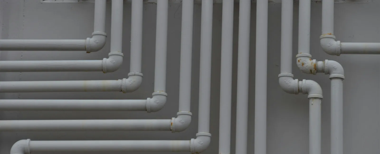 Network of white plumbing pipes with multiple joints mounted on a gray wall in a building.