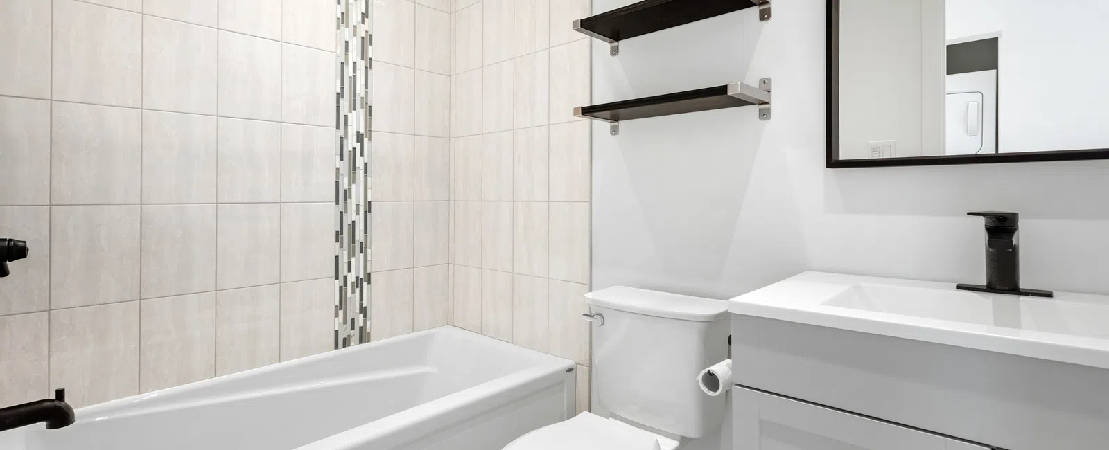 Modern bathroom with white bathtub, toilet, tiled walls, floating shelves, and vanity with black faucet and mirror.