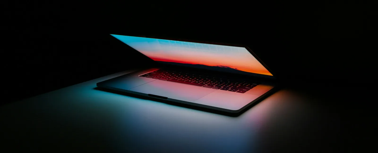 Partially open laptop glowing with colorful screen light in a dark room casting blue and red hues on surface.