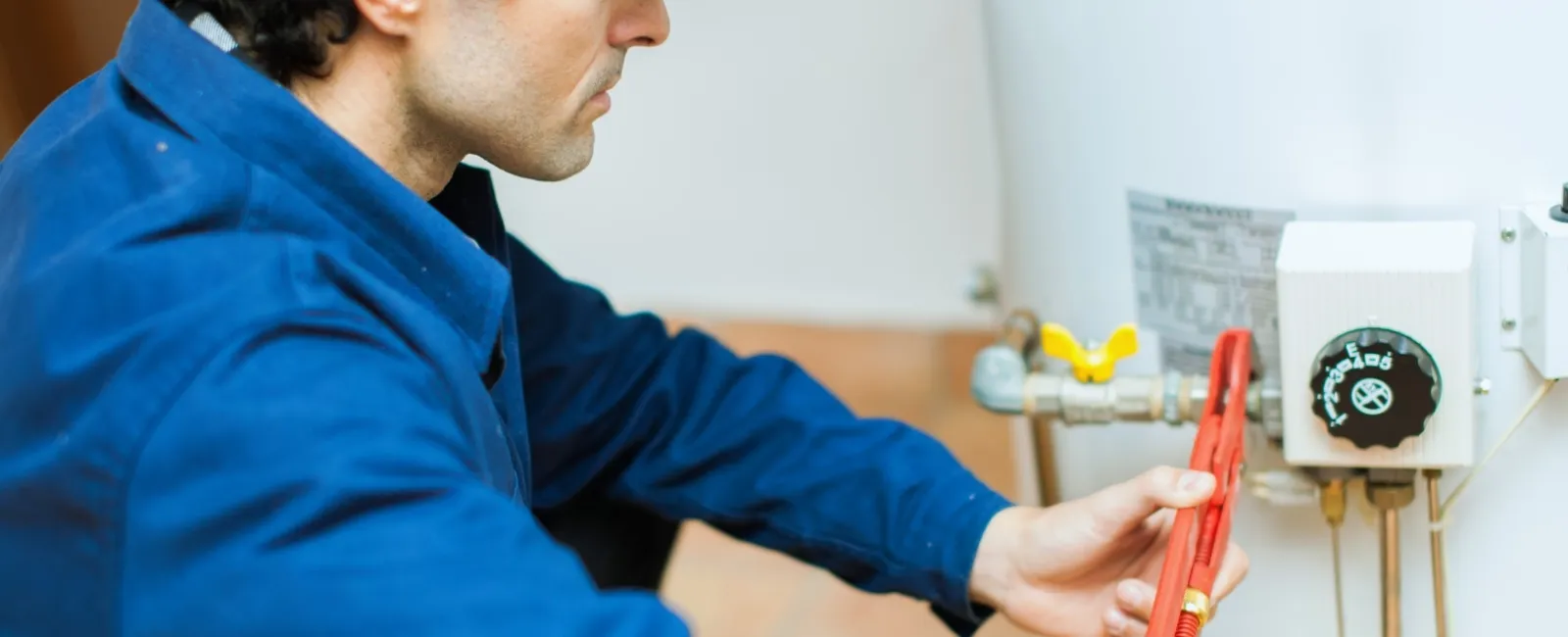 Technician in blue uniform using a wrench to repair a white water heater indoors.