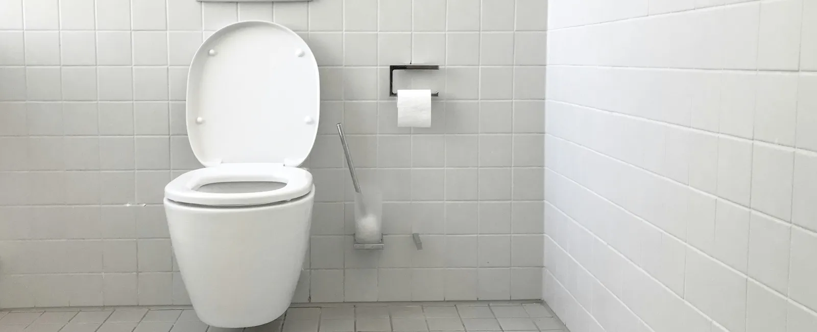 Modern white wall-mounted toilet with open lid in a clean tiled bathroom with toilet paper and brush.