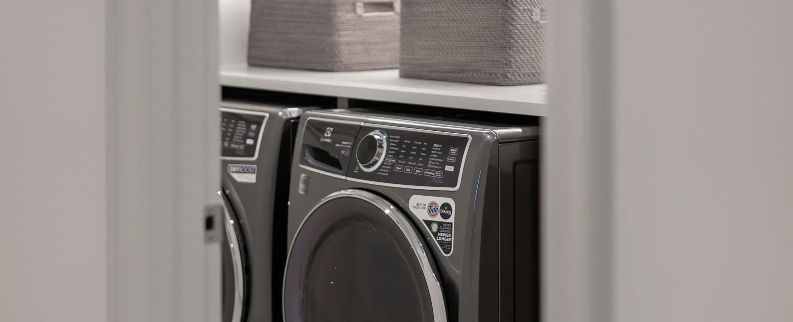 Modern laundry room with front-loading washer and dryer, gray cabinets, woven baskets, and herringbone tile floor.