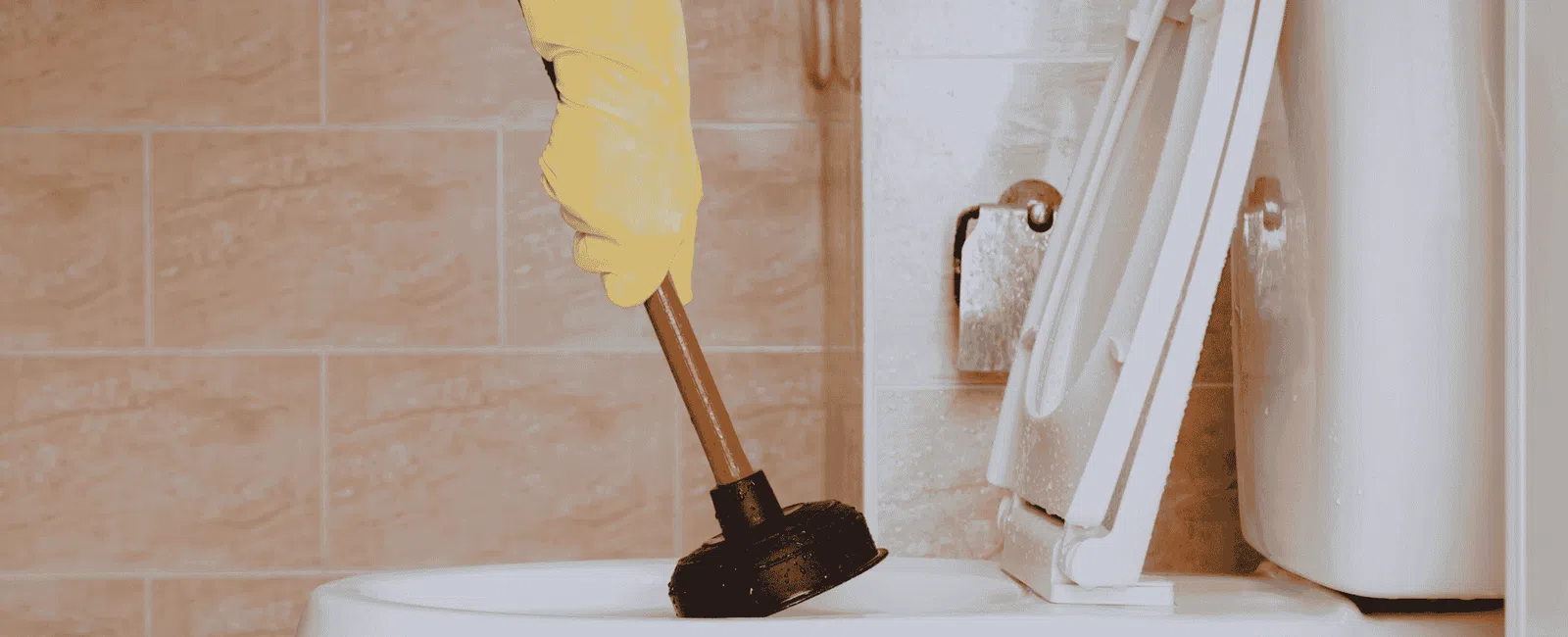 Hand with yellow glove using plunger to unclog white toilet in beige tiled bathroom.