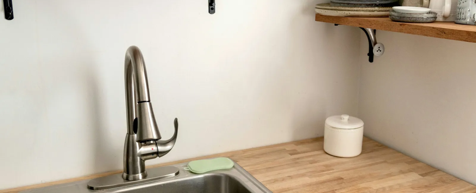 Modern kitchen corner with stainless steel sink, wooden countertop, open shelves with dishes and glasses.